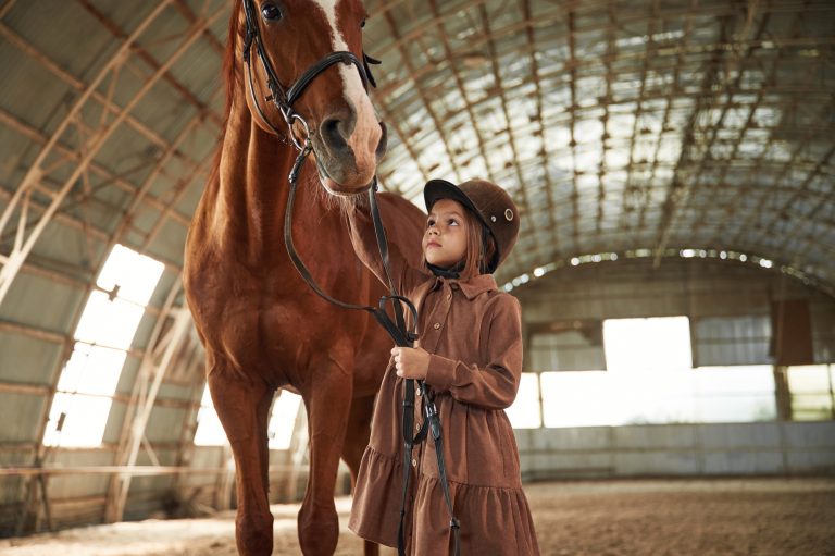 In jockey clothes. Cute little girl is with horse indoors